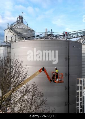 A male worker on an elevated work platform or cherry picker working at height on large industrial silos or storage tanks with copy space Stock Photo