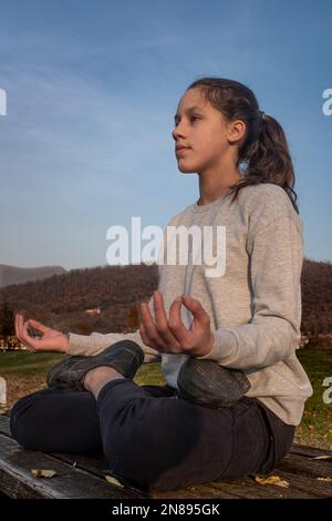 Teenage girl exercising outdoors and practicing yoga exercises in the park, sitting on a wooden table. Young woman finding her inner peace. Stock Photo