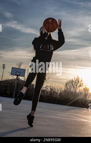 Front view of a teenage girl takes a shot on a basketball court, backlit sunset. Photograph capturing the lifting of feet off the ground. Stock Photo