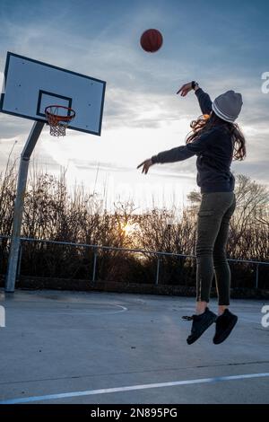 Rear view of a teenage girl, in mid-air, takes a shot on a basketball court, backlit sunset, vertical composition, copy space. Stock Photo