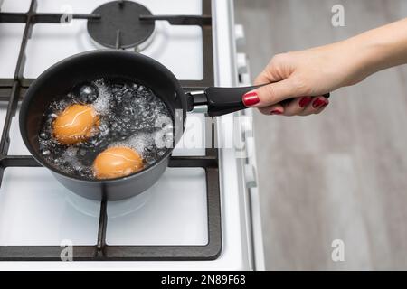 woman in the kitchen boils a chicken egg in water. man cooking an egg. chicken egg cooking. broken egg shell. Stock Photo
