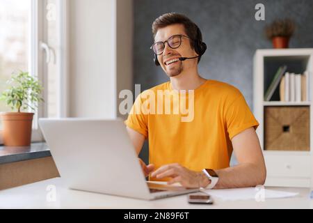Happy laughing man wearing headset communicate by conference video call with friends or family. Freelancer working remotely from home with laptop, smiling. Student has online class webinar course Stock Photo