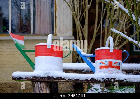 old watering cans painted and decorated in colours with flags of uk and hungary on snow covered garden bench Stock Photo