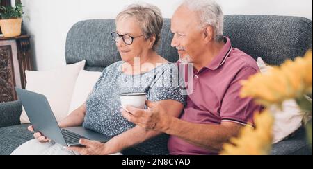 Happy couple of senior man and woman at home using laptop together in indoor leisure activity. Technology and mature people. Wireless computer connect Stock Photo