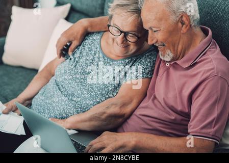 One happy and jyful old senior couple at home enjoying internet and techology leisure actiity togethe rusing laptop sitting on sofa. Modern senior peo Stock Photo
