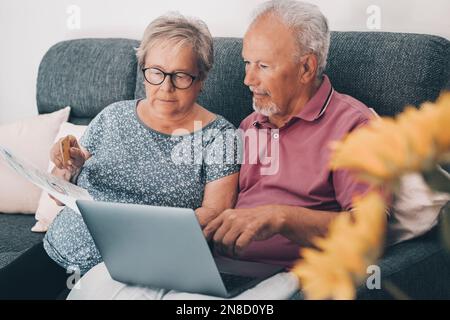 One old senior couple at home paying bills online with web banking app and checking the amounts on documents. Elderly and modern technology real life. Stock Photo