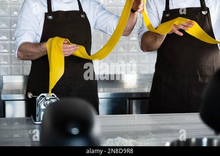Fresh pasta and pasta machine. Italian traditional tagliatelle. Young men in apron holding the dough for pasta at kitchen. Stock Photo