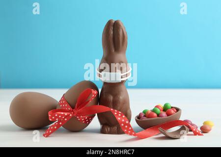 Chocolate bunny with protective mask and eggs on white wooden table. Easter holiday during COVID-19 quarantine Stock Photo