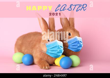 Text Happy Easter 2021 and cute bunnies in protective masks on pink background. Holiday during Covid-19 pandemic Stock Photo