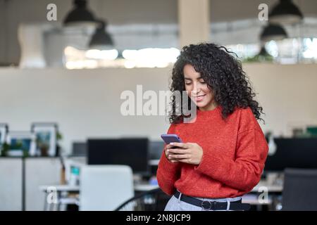 Smiling young latin business woman using phone standing in office. Stock Photo