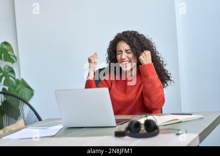 Young excited latin woman raising hands winning online getting email on laptop. Stock Photo