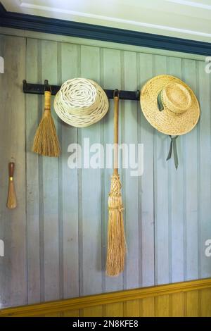 Vintage weaved straw hats and broomsticks hangimg on light green painted wood plank wall in entryway inside old 1809 French regime cottage style home. Stock Photo