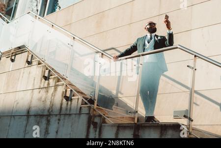 A well-dressed bald bearded black man wearing a suit and sunglasses, looking at his cell phone while standing on the stairway with a seamless glass ra Stock Photo