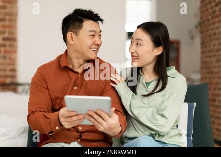 Happy asian middle aged man and young woman sitting on sofa and using digital tablet, looking at each other and smiling Stock Photo