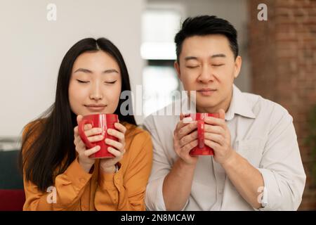 Calm asian young woman and middle aged man smelling fresh aromatic morning coffee with closed eyes Stock Photo