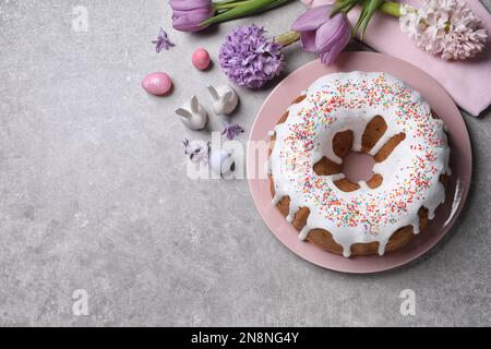 Glazed Easter cake with sprinkles, painted eggs and flowers on grey table, flat lay. Space for text Stock Photo