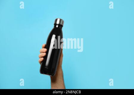 Man holding black thermos bottle on light blue background, closeup. Space for text Stock Photo
