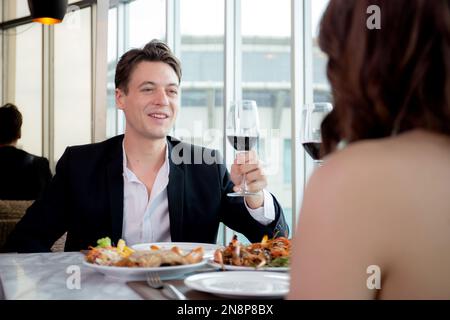 Happy young couple holding with glass of wine together in anniversary at restaurant for celebration, man and woman dinning while date with romance and Stock Photo