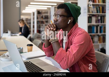Thoughtful black male student wearing wireless earbuds sitting at desk with laptop in library Stock Photo