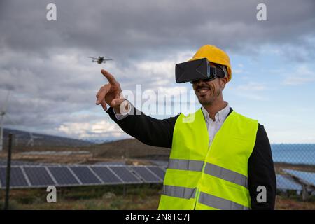 An engineer pilots a drone wearing VR headsets in a field of solar panels and wind turbines. Stock Photo