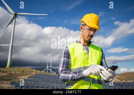 An engineer in a field of wind turbines and solar panels works using his phone, renewable energy Stock Photo