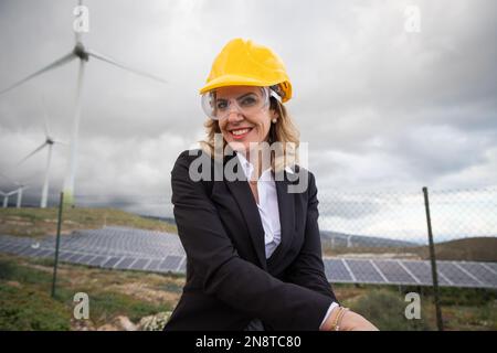 Portrait of a smiling female engineer in the photovoltaic power station with wind turbines. Stock Photo