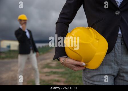 Portrait of a helmet held in hand by a female engineer with her colleague out of focus behind. Stock Photo