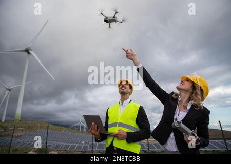 Two engineers at work fly a drone and use the laptop in a solar farm with wind turbines Stock Photo