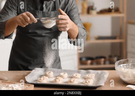 Male baker sprinkling raw buns with flour at table in kitchen, closeup Stock Photo