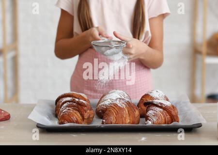 Little baker sprinkling sugar powder onto croissants in kitchen, closeup Stock Photo