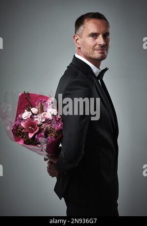 Surprise, portrait and man with flowers for a date isolated on a grey studio background. Classy, gift and mature person in a suit with a bouquet of Stock Photo