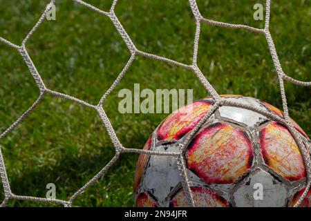 old soccer ball in the net on the background of grass soccer field. Summer sunny day. Stock Photo