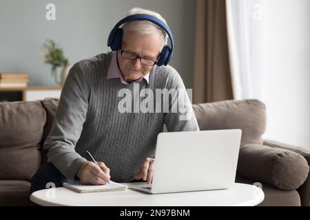 Focused senior retired man in wireless earphones writing in copybook Stock Photo