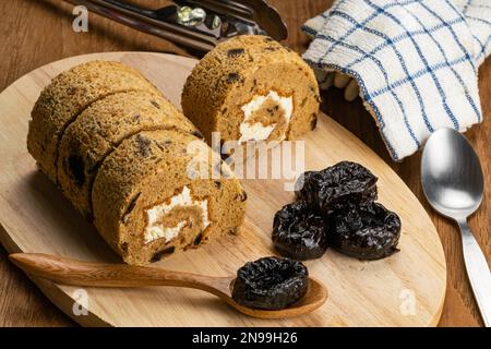 Hogh angle view of homemade sponge cake roll and dried pitted prune fruit on wooden board with metal spoon, metal tongs and tablecloth on wooden table Stock Photo