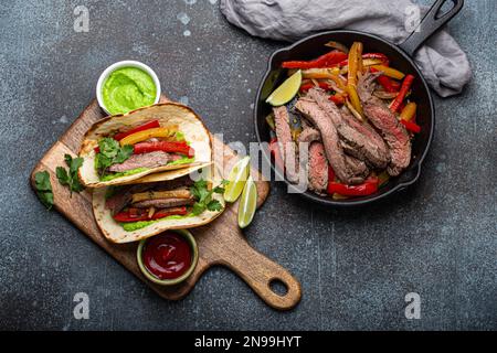 Process of making traditional Mexican dish Beef fajitas tacos served on wooden cutting board with tomato salsa and guacamole on rustic stone Stock Photo