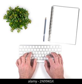 top view of person typing on wireless computer keyboard on white desk with potted plant and notepad with pencil Stock Photo