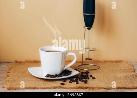 White cup of hot coffee with beans on sackcloth napkin and milk frother on wooden table against beige wall. Selective focus. Stock Photo