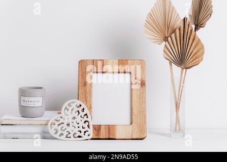 Mockup wooden picture frame, books, aroma candle, carved decor heart, fan leaves in vase on white table. Copy space. Stock Photo