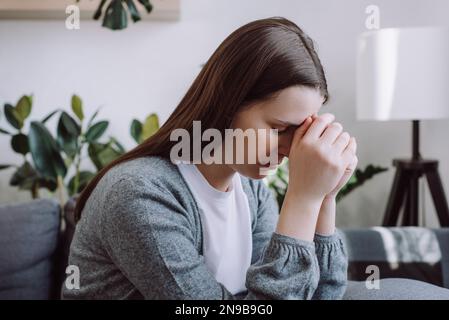 Close up of concerned sad young woman sitting on couch at home thinking of problem looks in distance feels depressed due loneliness, inner emptiness, Stock Photo