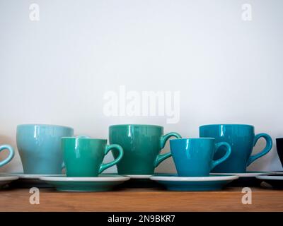 Set of big and small blue green ton of coffee mugs, ceramic cups with saucers stand in a row on wooden shelf isolated on white background with copy sp Stock Photo