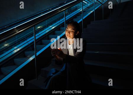 Young asian woman using mobile phone while sitting on the stairs next to escalator Stock Photo