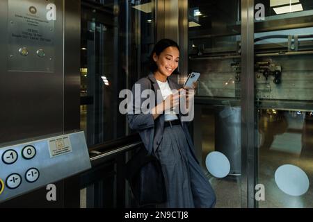Young asian woman smiling and using smart phone while standing in elevator Stock Photo