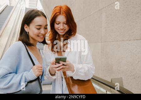 Young multinational women smiling and using cellphone while standing indoors Stock Photo