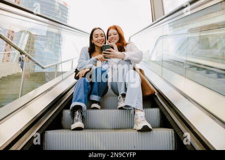 Young multinational women smiling and using cellphone while sitting on escalator Stock Photo