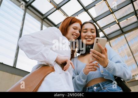 Young multinational women smiling and using cellphone while standing indoors Stock Photo