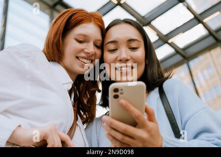 Young multinational women smiling and using cellphone while standing indoors Stock Photo