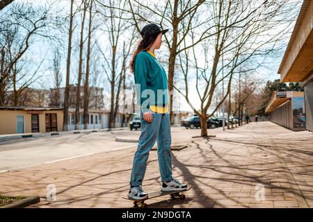 A young Caucasian hipster woman rides down the street on a skateboard. In the background, an alley. Concept of sports lifestyle and street culture. Stock Photo