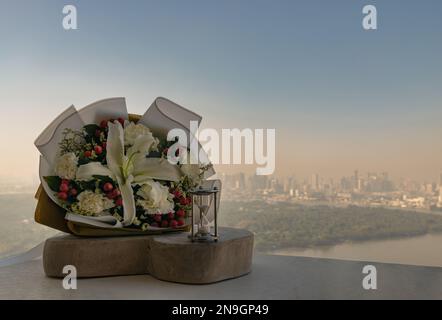 Hourglass or Sandglass is next to Bouquet of flowers placed on the timber outside the balcony with the city view background. Symbol of Romantic foreve Stock Photo
