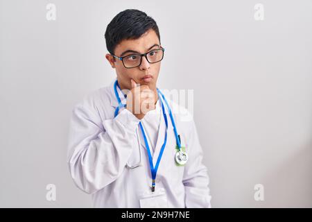 Young hispanic man with down syndrome wearing doctor uniform and stethoscope serious face thinking about question with hand on chin, thoughtful about Stock Photo