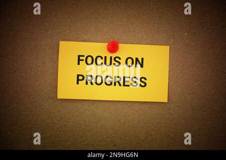 A yellow paper note with the phrase Focus On Progress on it pinned to a cardboard background. Close up. Stock Photo